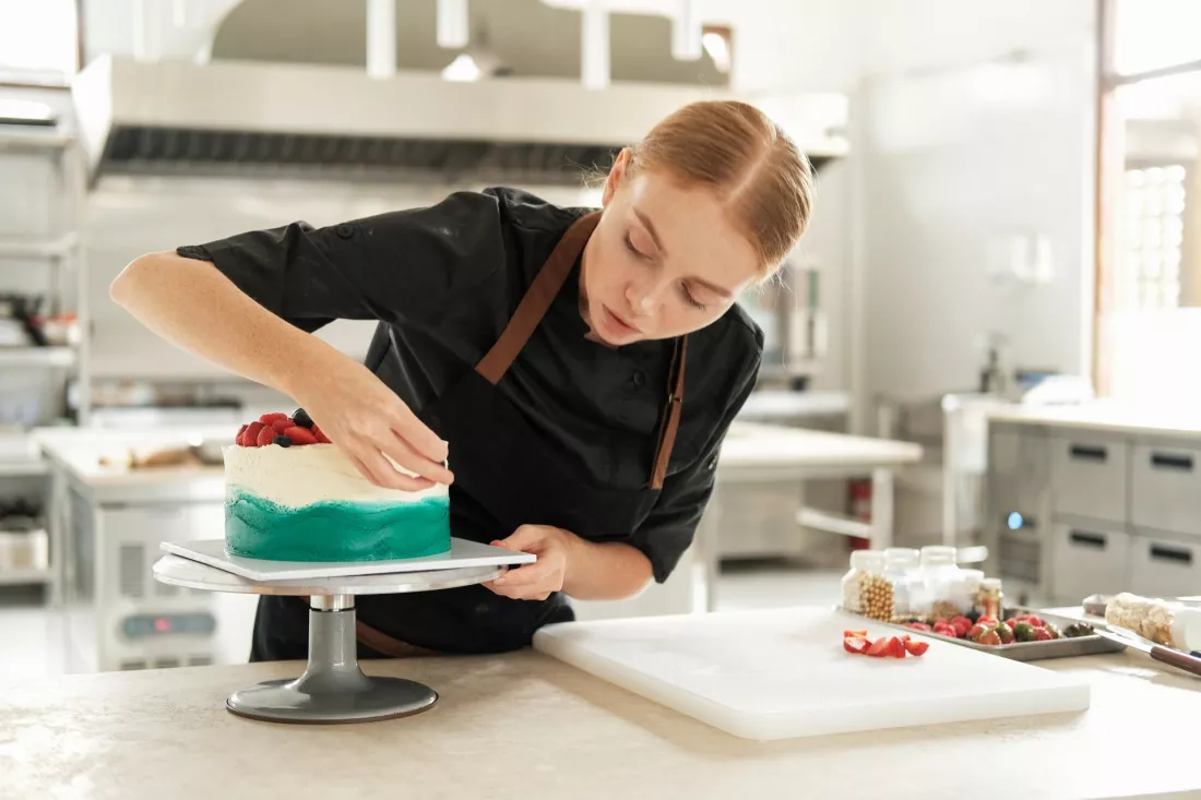 Młoda cukierniczka dekoruje tort w pracowni cukierniczej.